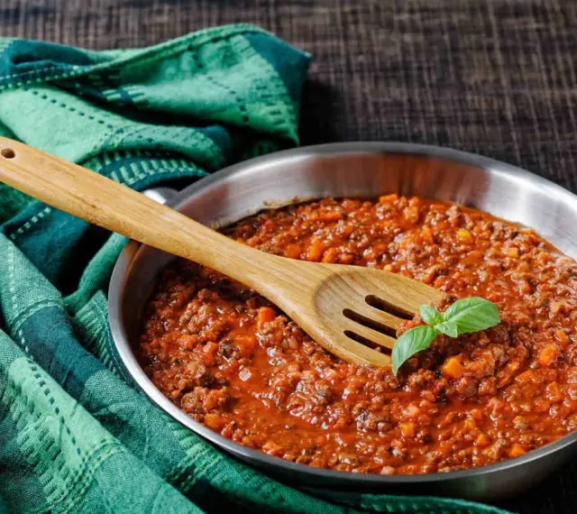 Un saporito ragù di cinghiale in padella, pronto per condire la pasta. Un cucchiaio di legno e una fogliolina di basilico completano la scena.
