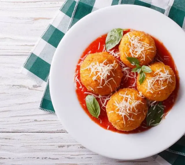 Quattro arancini di riso dorati, cosparsi di parmigiano e serviti su un letto di salsa di pomodoro, guarniti con basilico fresco.