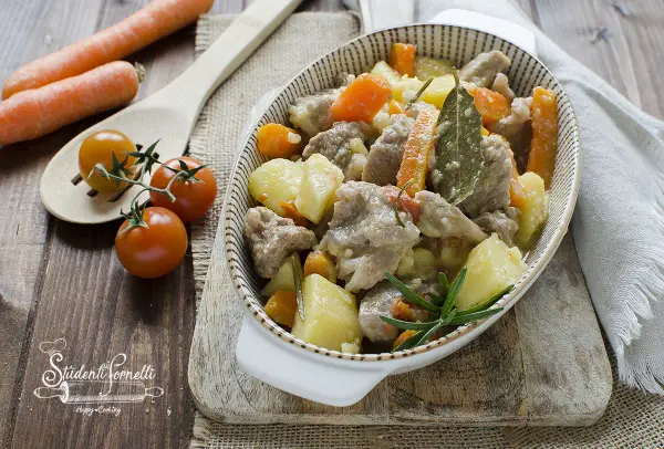 Un invitante spezzatino in bianco con patate e carote, guarnito con rosmarino e alloro. Accanto, pomodorini e carote fresche.