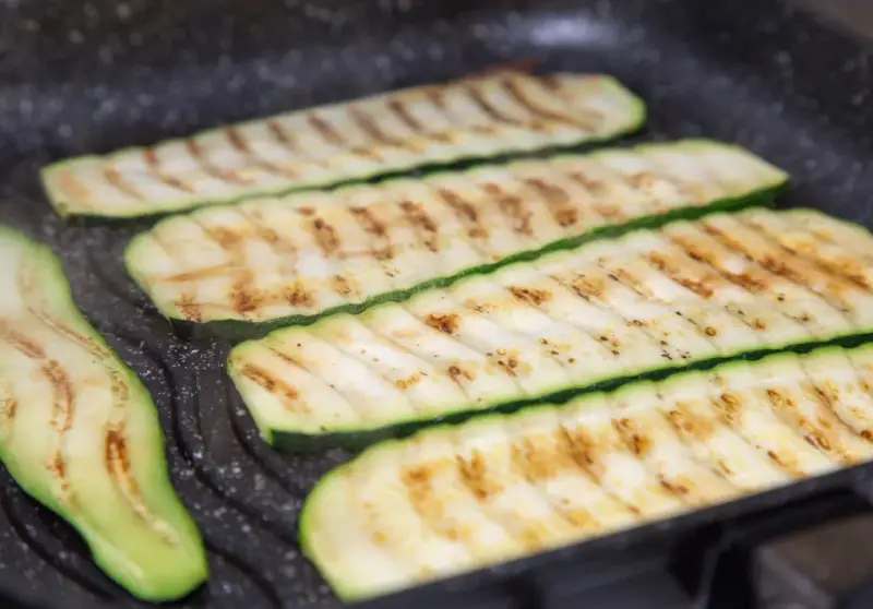 Fette di zucchine grigliate che cuociono su una piastra, con segni di grigliatura dorati e invitanti.